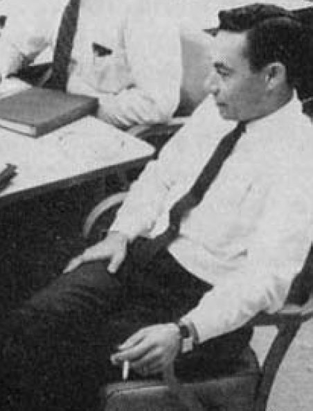 Mohamed Atalla seated at the corner of a table smoking a cigarette during a meeting. Other people are present but their faces aren't visible.